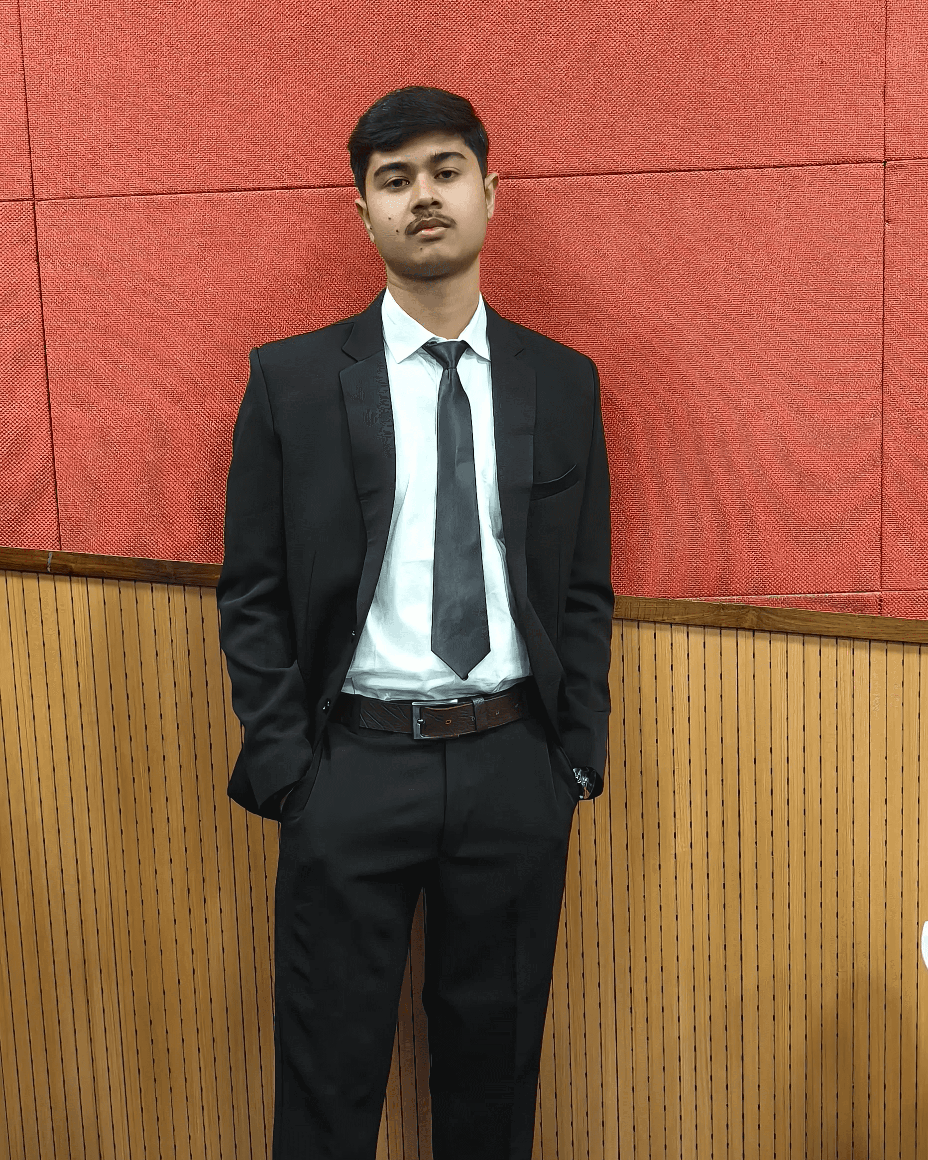 Ayush Singh in a suit, standing against a red and wooden panelled wall